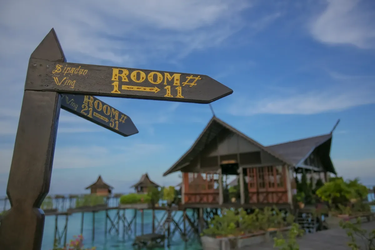 Wooden signage directing rooms at a tropical beach resort with clear blue skies and stilt houses.